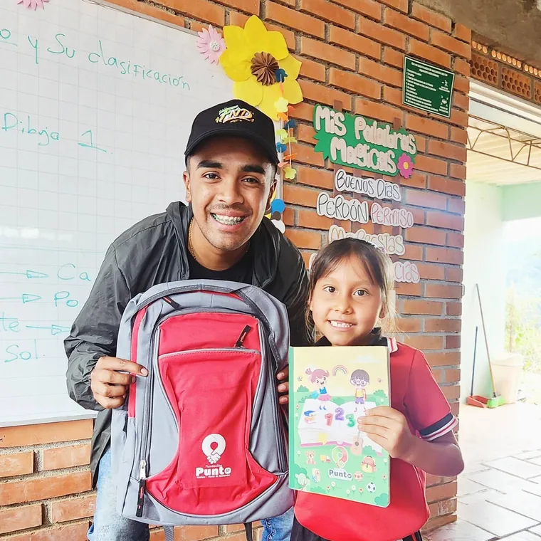 Voluntario y niña muestran un morral y un cuaderno entregados por la fundación
